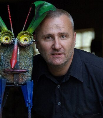Ein Nahaufnahme-Portrait des Künstlers Joest Leopold mit kurz rasierter Frisur und schwarzem Hemd. Er blickt direkt in die Kamera. Links neben ihm steht eine große, farbenfrohe Upcycling-Skulptur, die einen stilisierten Charakter mit großen Augen aus runden Metallteilen, einer roten Nase und grünen Blättern als Kopfschmuck darstellt. Der Hintergrund ist unscharf und dunkel.