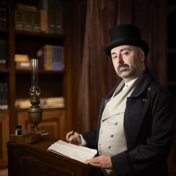 Darsteller in 19. Jahrhundert-Kleidung vor Bücherregalen, mit Schreibutensilien und Laterne auf dem Pult.