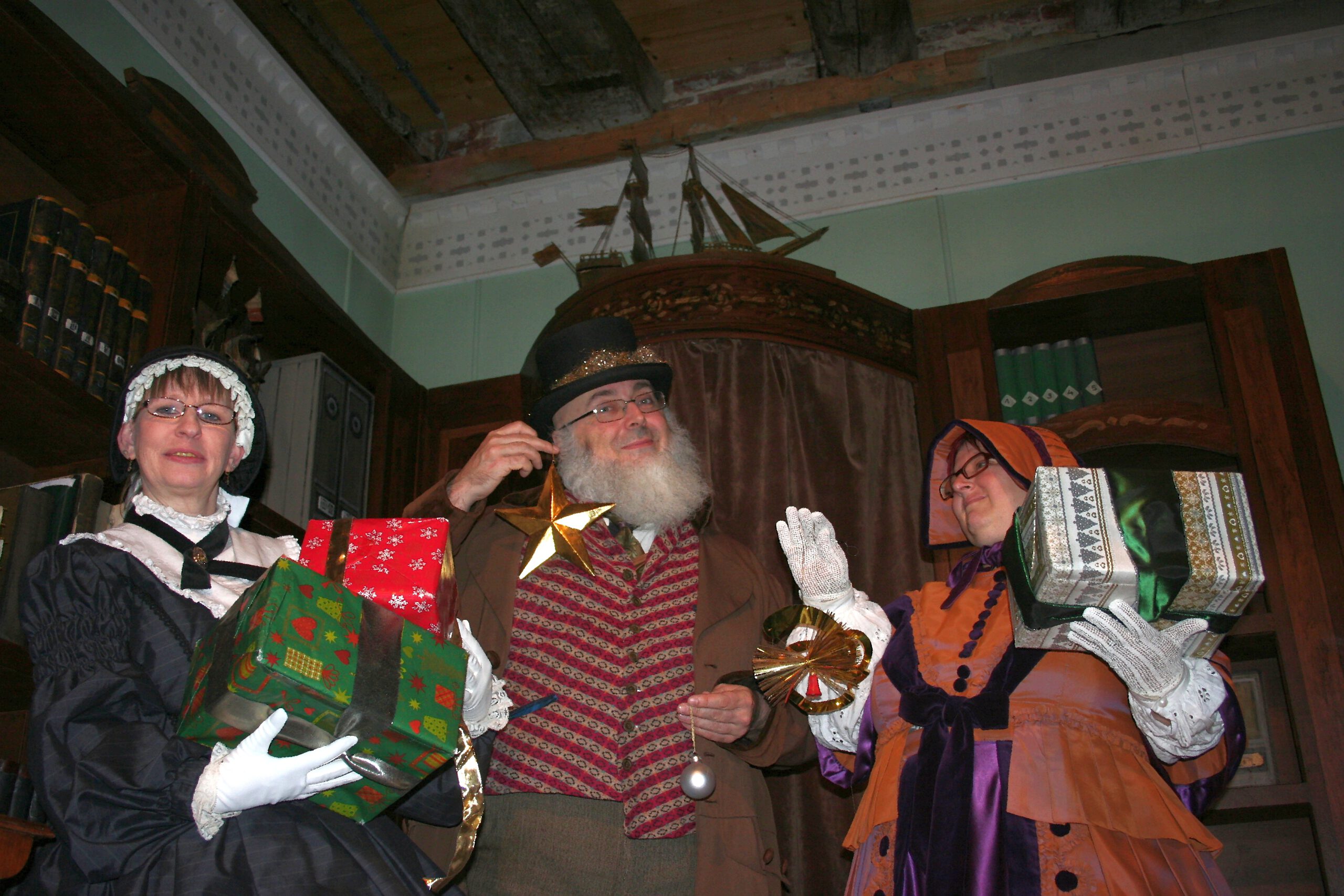 Drei Personen in historischen Kostümen des 19. Jahrhunderts stehen in einem holzgetäfelten Raum und halten festlich verpackte Geschenke. In der Mitte steht ein Mann mit weißem Bart, Zylinder und Stern in der Hand, flankiert von zwei Frauen in viktorianischer Kleidung mit Hauben und bunten Kleidern. Die Szene vermittelt eine weihnachtliche, nostalgische Stimmung.