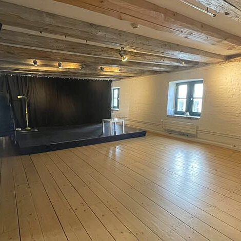 Bühne Ein heller Veranstaltungsraum mit rustikalem Charme, geprägt durch helle Holzdielenböden und eine niedrige Decke mit schweren Sichtbalken aus dunklem Holz. Im hinteren Teil befindet sich eine niedrige, schwarz verkleidete Bühne vor einem schwarzen Vorhang, auf der ein kleiner weißer Beistelltisch steht. Die weißen Backsteinwände werden durch kleine Fenster unterbrochen, die natürliches Tageslicht in den Raum lassen. Links im Vordergrund sind Ansätze von Bühnenequipment und schwarzen Vorhängen zu sehen.