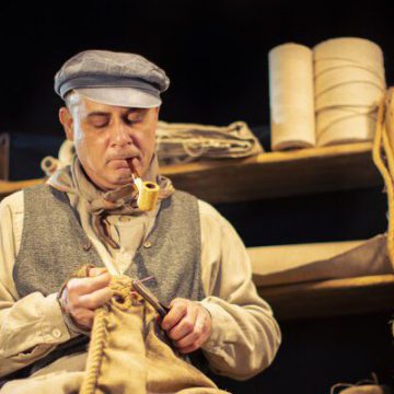 Darsteller in traditioneller Hafenarbeiter-Kleidung stopft einen Sack, unter einer Laterne, mit Spulen und Tauwerk um sich herum.